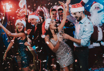 Happy Young People Dancing on New Year Party. Happy Young People Dancing on New Year Party. Santa Claus. People in Red Caps. Happy New Year Concept. Glass of Champagne. Celebrating of New Year. Young Woman in Dress. Men in Suits.